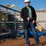 Place of Worship building in process. Pastor with a hard hat stands in front of the building with a construction worker behind on a lift
